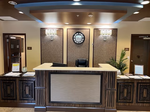 Reception desk area with a large wooden counter, two computer monitors, a black office chair, decorative wall sconces, a round wall clock, and potted plants. There are two doors on either side of the reception desk with signage and papers on the counter.
