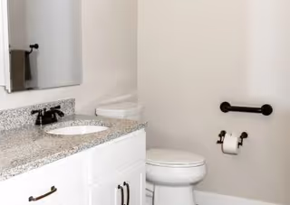 A clean and simple bathroom featuring a white toilet, a white vanity with a granite countertop and an undermount sink, a large mirror above the sink, a towel hanging on a black towel holder, and a black toilet paper holder mounted on the wall.