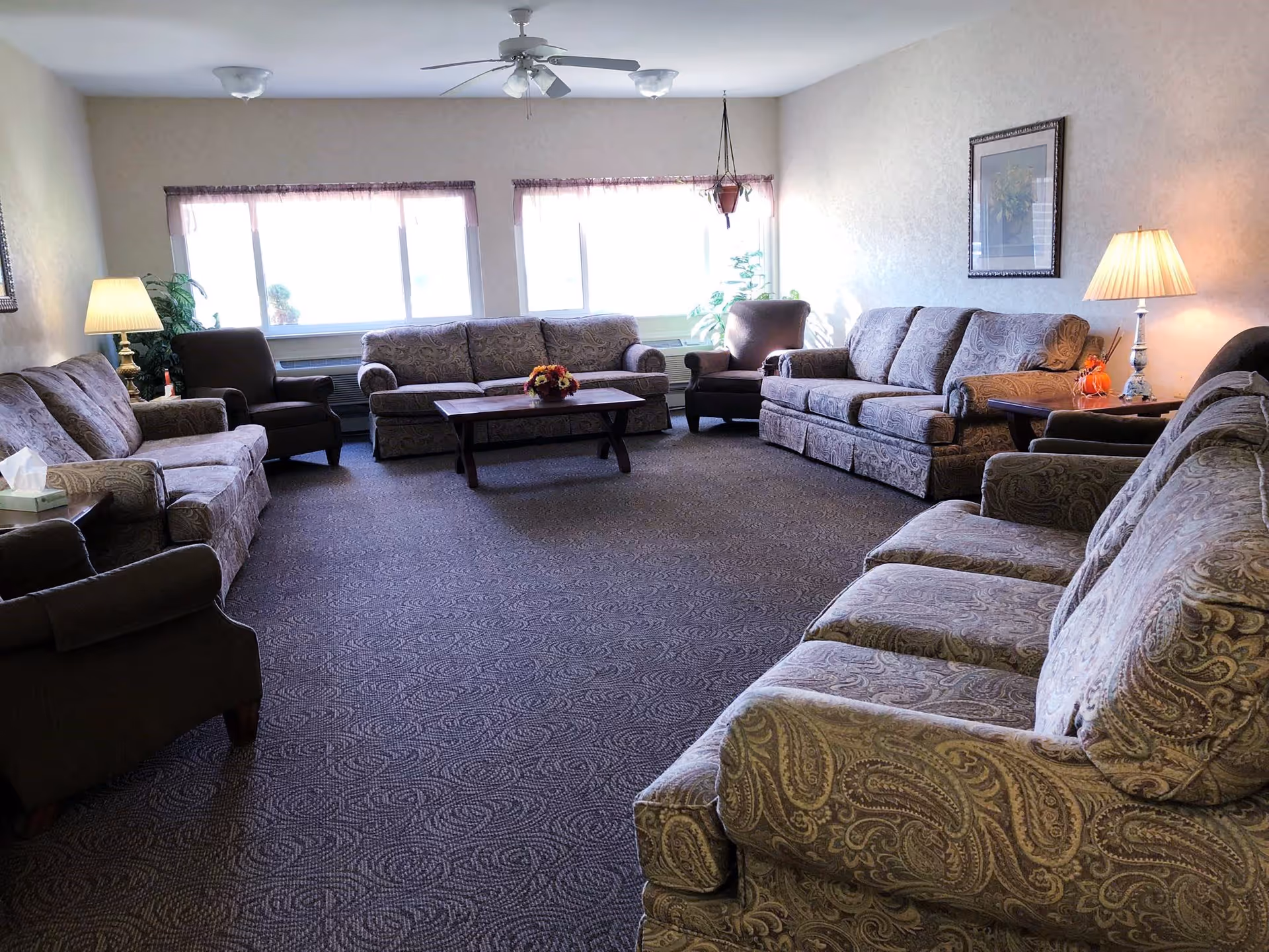 A spacious living room with multiple patterned sofas and armchairs arranged around a wooden coffee table. The room has large windows letting in natural light, a ceiling fan with lights, table lamps, framed artwork on the wall, and some potted plants.