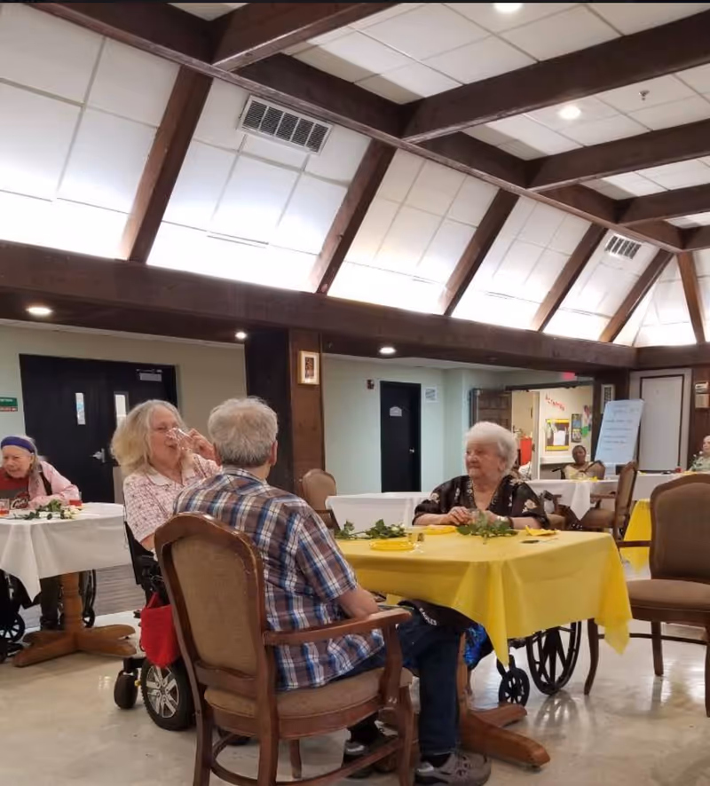 Several elderly people sitting at tables in a communal dining or activity room with yellow and white tablecloths. The room has a high ceiling with exposed wooden beams and bright lighting. Some individuals are in wheelchairs, and the atmosphere appears calm and social.