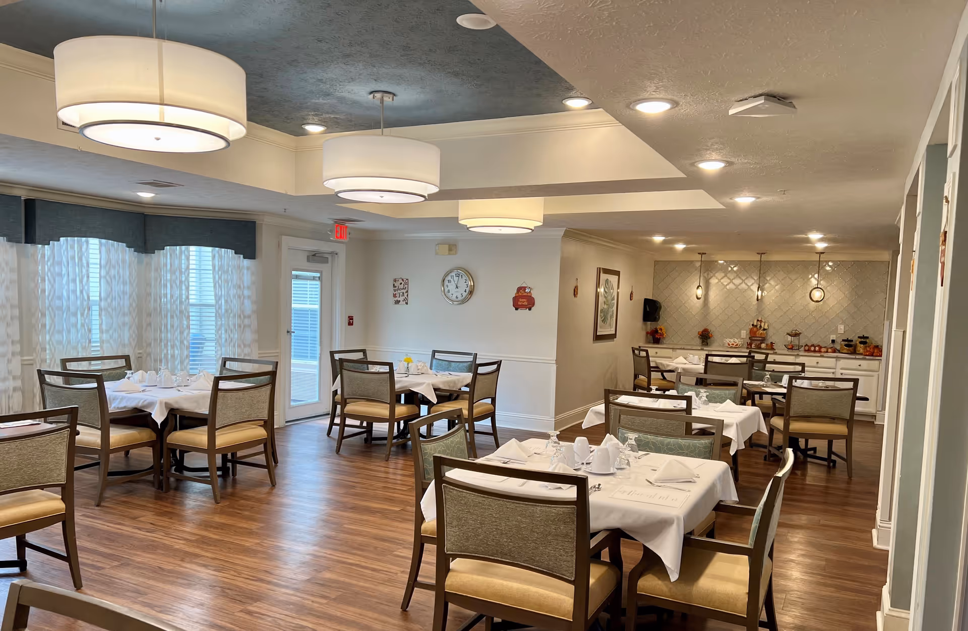 A dining room in a senior living facility with several tables covered in white tablecloths, each set with napkins, glasses, and cups. The room has wooden floors, large windows with sheer curtains, modern ceiling lights, and a buffet area with food and decorations in the background.