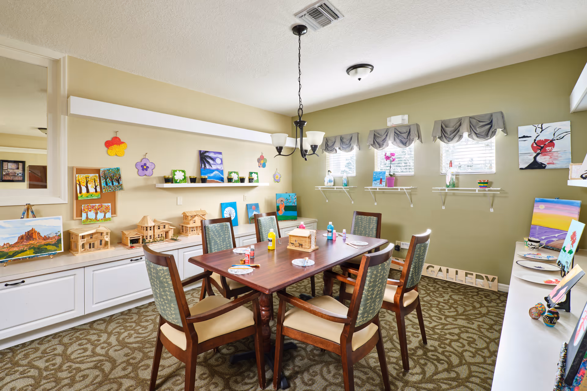 A bright room with a wooden table surrounded by six chairs. The table has art supplies including paint bottles and plates. The walls are decorated with various paintings, small wooden house models, and colorful flower-shaped wall hangings. There are three small windows with gray valances letting in natural light. The carpet has a patterned design and the room has a cozy, creative atmosphere.