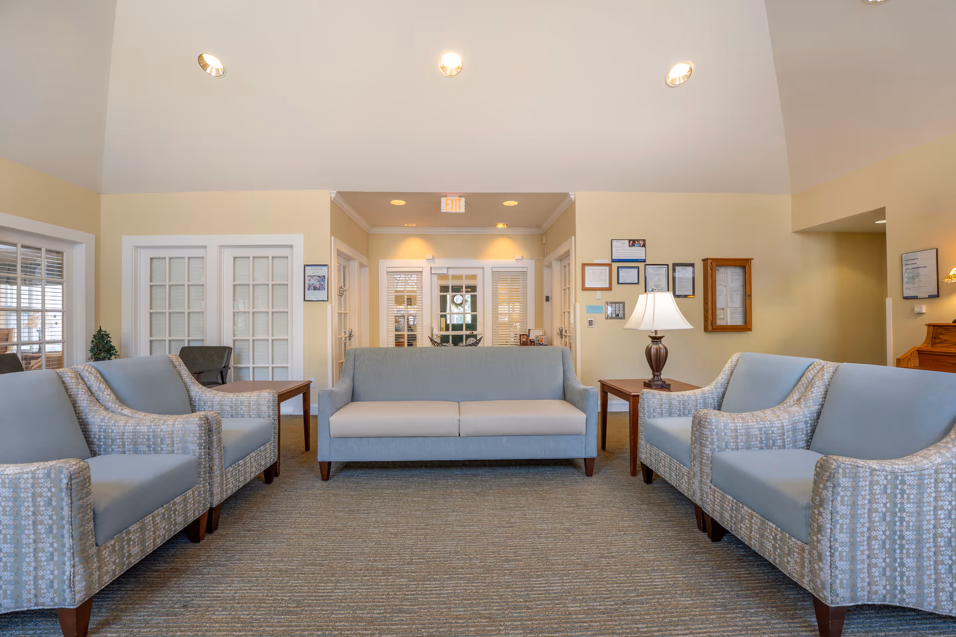A well-lit seating area in a senior living facility with a central light blue sofa flanked by two matching armchairs on each side. The walls are painted light yellow, and there are framed certificates and a lamp on a wooden side table to the right. French doors and windows with white frames are visible in the background.
