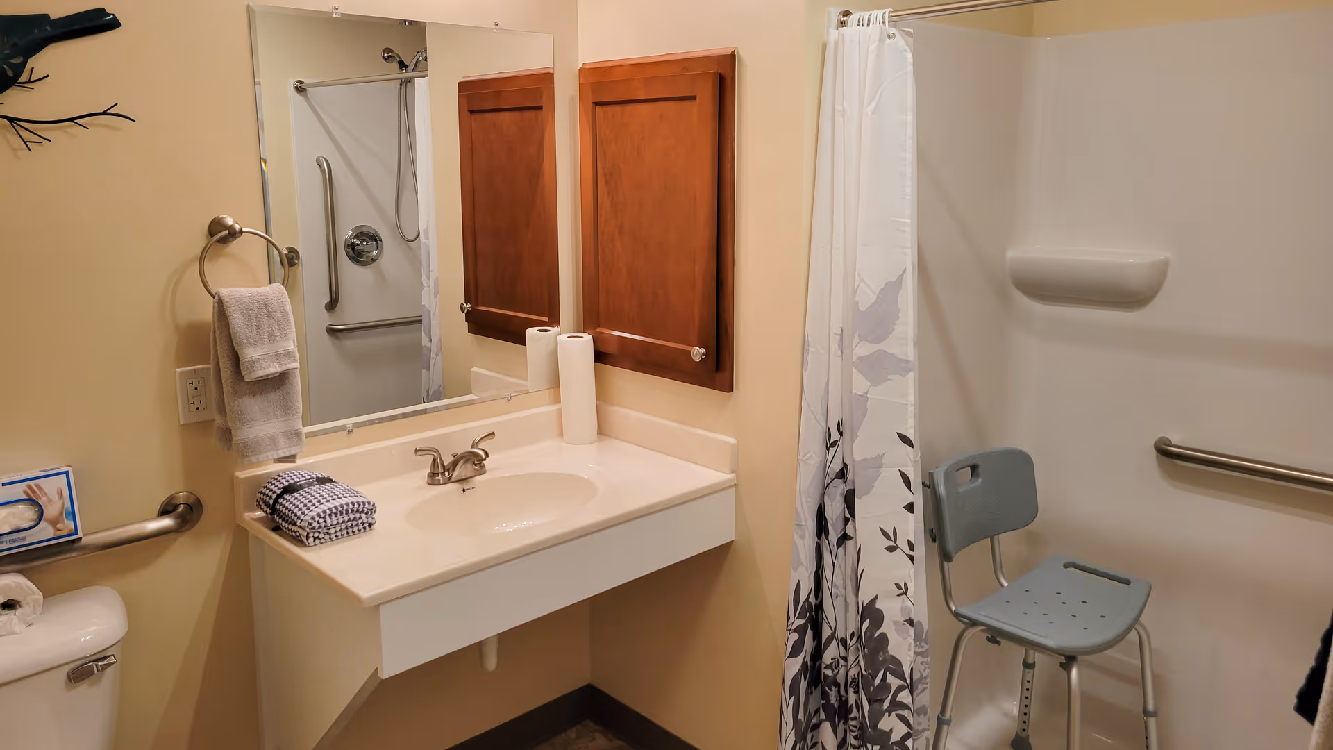 Accessible bathroom with a sink and mirror, wooden medicine cabinets, grab bars, a shower with curtain and a shower chair.
