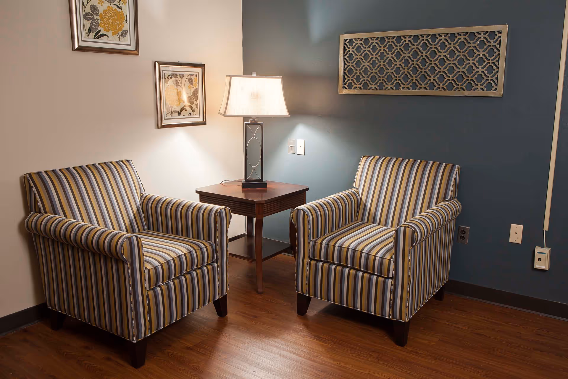 Two striped armchairs with yellow, gray, and white stripes are positioned on either side of a wooden side table with a lamp on it. The room has wooden flooring, a blue accent wall with a decorative wooden panel, and two framed floral artworks on a white wall.