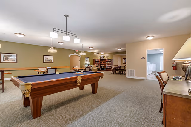 A spacious common area in a senior living facility featuring a pool table with balls set up, several tables and chairs, bookshelves filled with books, and a seating area with armchairs. The room has beige walls, carpeted floors, and ceiling lights providing warm illumination.