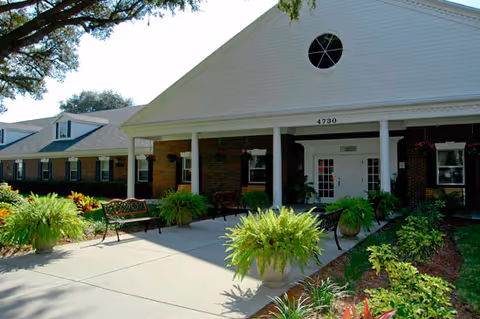 Exterior view of a single-story brick building with a white triangular pediment and columns at the entrance. The entrance area has benches, potted plants, and hanging flower baskets. The building number 4730 is visible above the door. There are trees and landscaped greenery around the walkway leading to the entrance.
