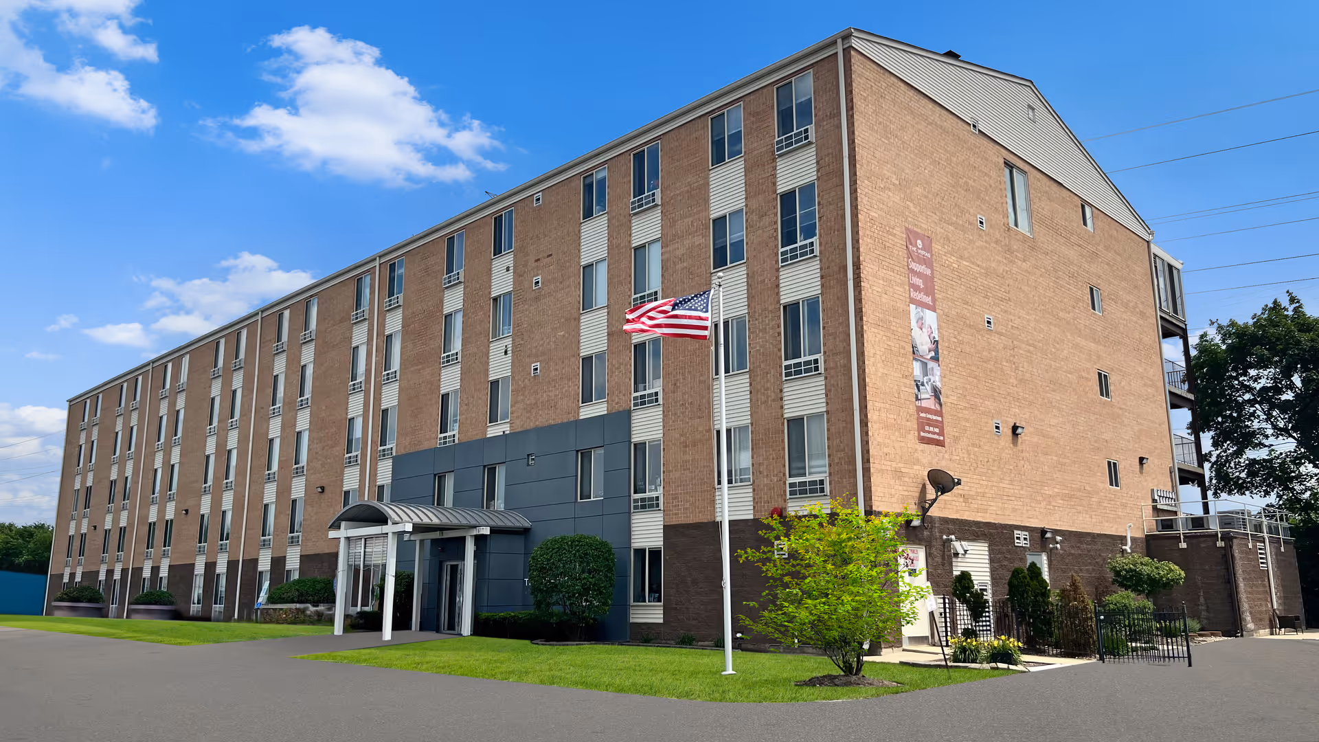 Exterior view of a multi-story brick building with numerous windows, a main entrance with a small canopy, an American flag on a flagpole, and some landscaping including bushes and a small tree under a partly cloudy blue sky.