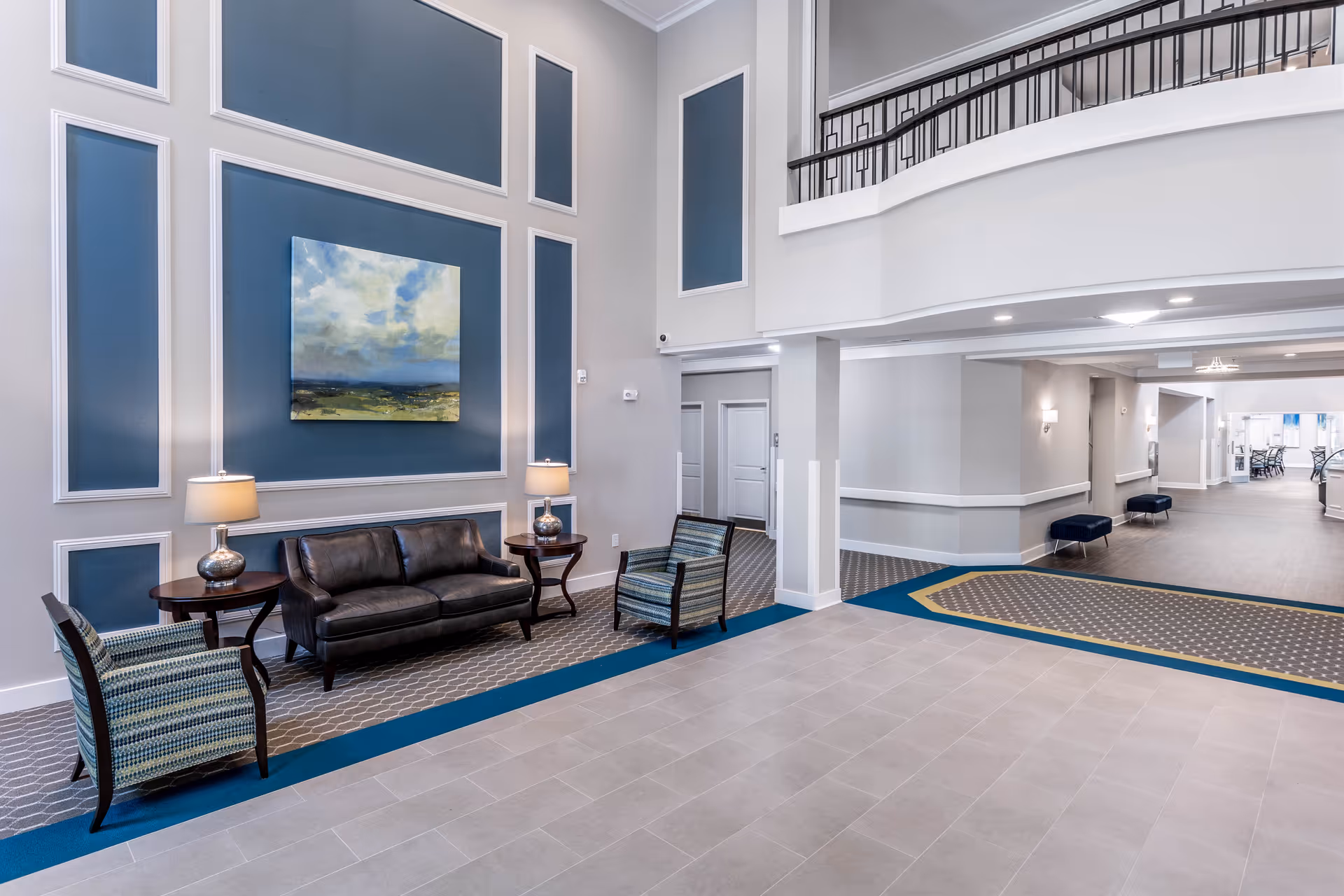 Spacious senior living facility lobby with seating area, artwork on the wall, and a second-floor balcony.