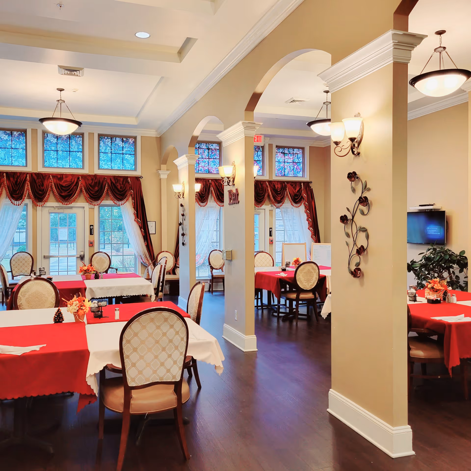 A bright and elegant dining room in a senior living facility with tables covered in red and white tablecloths, surrounded by upholstered chairs. The room features arched openings, decorative wall sconces, large windows with red valances and sheer curtains, and dark wood flooring.