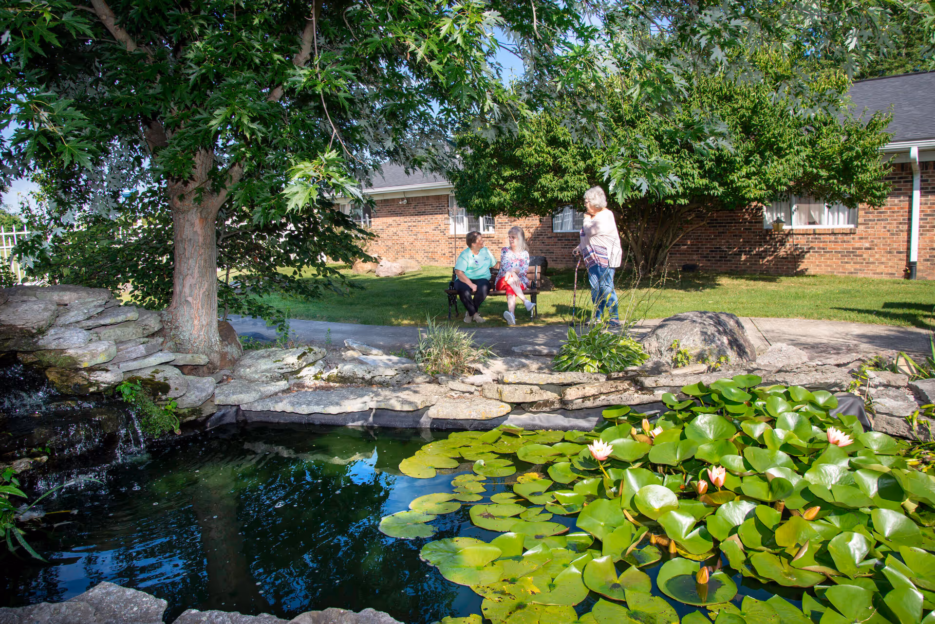 A peaceful outdoor garden area at a senior living facility with a small pond filled with lily pads and blooming flowers. Three elderly women are seen enjoying the sunny day; two are seated on a bench under a tree, and one is standing nearby with a cane. A brick building is visible in the background.