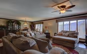 A spacious living room with multiple beige sofas and a large ottoman in the center. The room features a ceiling fan with lights, large windows with blinds allowing natural light, and decorative plants and furniture along the walls.