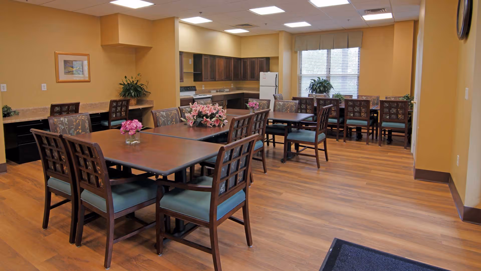 A dining area in a senior living facility with multiple wooden tables and chairs arranged neatly. The room has wood flooring, beige walls, and a kitchen area with dark cabinets, a stove, and a refrigerator in the background. There are decorative plants and flowers on the tables and counters, and a large window letting in natural light.