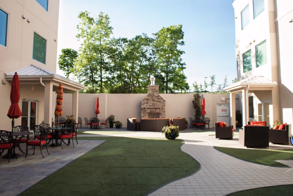Outdoor courtyard area at Livingston Post Acute Care with patio furniture including tables, chairs with red cushions, and umbrellas. There is a stone fireplace against a beige wall, surrounded by seating. Trees are visible beyond the wall under a clear blue sky.