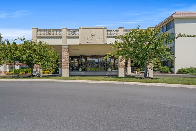 Front exterior view of a senior living facility named Spring Hills Assisted Living, showing the entrance with glass doors, stone pillars, and trees on either side under a clear blue sky.