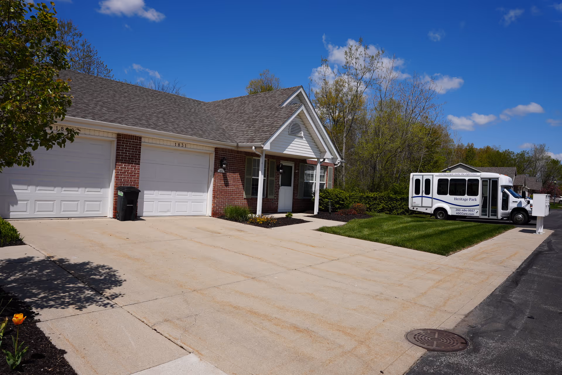 Exterior view of a single-story brick building with two white garage doors and a small covered entrance. A white shuttle bus labeled 'Heritage Park' is parked on the grass beside the building. The sky is clear with a few clouds, and there are trees and shrubs around the building.
