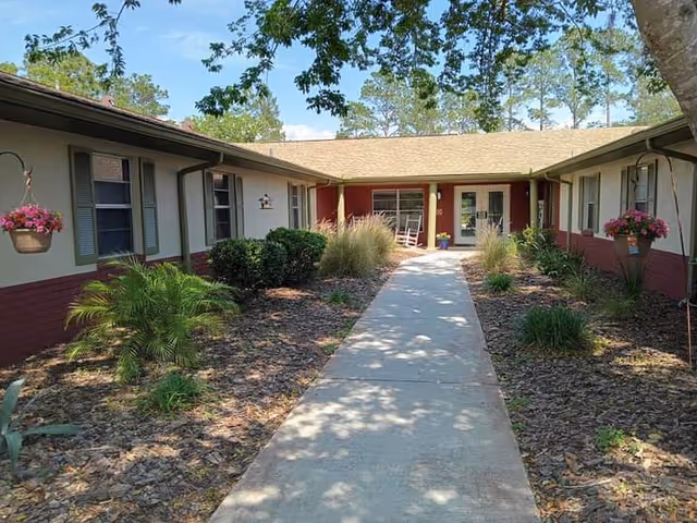 Concrete walkway flanked by landscaped beds and hanging flower baskets leading to the entrance of a single-story residential building.
