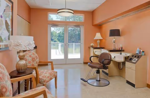 A small salon-style interior with a styling chair and wash sink, patterned waiting chairs, lamps, and double doors leading outside.