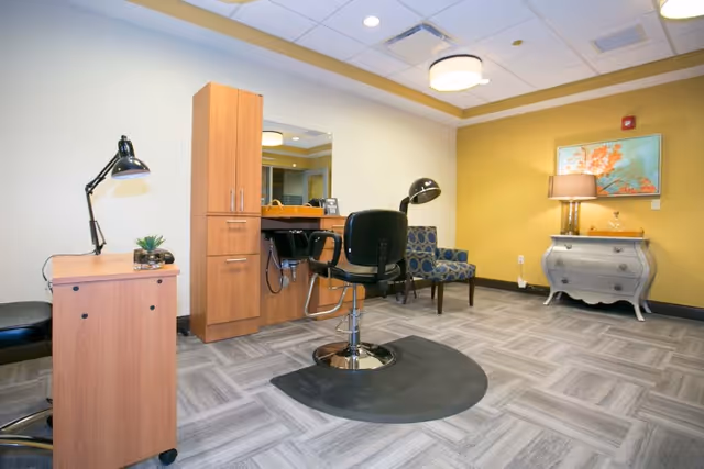 A bright salon-style room with a styling chair at a mirror station, cabinets, lamps and additional seating.