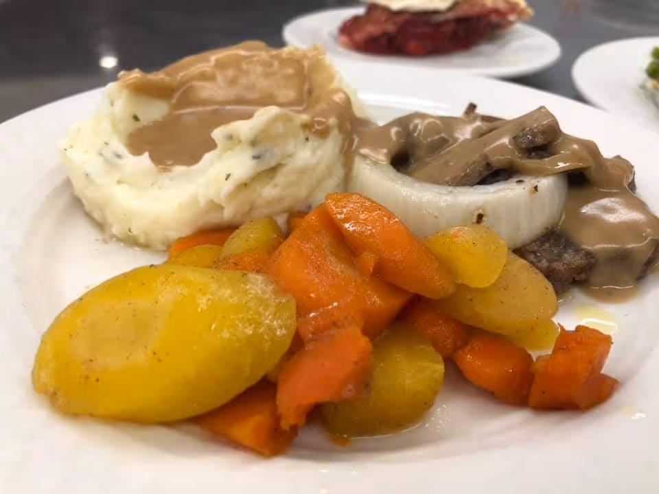 A white plate with a serving of mashed potatoes topped with brown gravy, cooked carrots and yellow squash, and a piece of meat with mushroom gravy and a slice of onion.