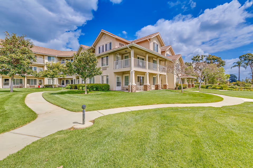 Exterior view of a senior living facility building with beige walls and a red-tiled roof, surrounded by a well-maintained green lawn, trees, and a curved concrete walkway under a partly cloudy blue sky.