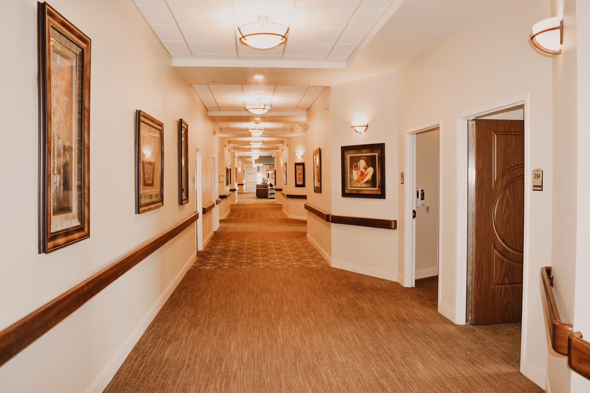 A well-lit, long hallway in a senior living facility with beige walls and carpeted floor. The hallway features wooden handrails on both sides and framed artwork hanging on the walls. Several doorways lead to rooms, one of which has the number 234 on the door frame. Ceiling lights and wall sconces provide warm lighting throughout the corridor.