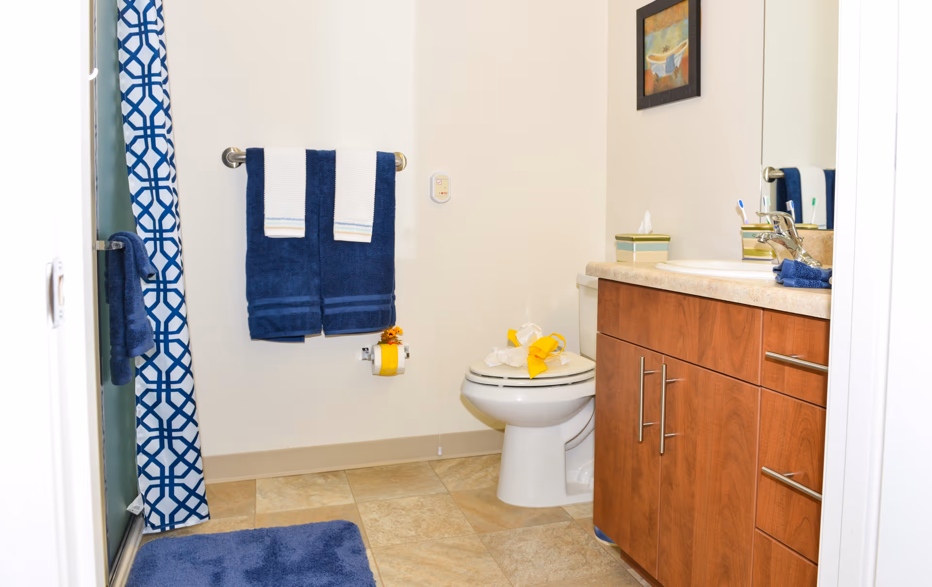 A clean bathroom with beige tiled floor, a wooden vanity with a sink and mirror, a toilet with a yellow decoration on the lid, a towel rack holding two navy blue towels with white accents, a blue and white patterned shower curtain, and a blue bath mat on the floor.