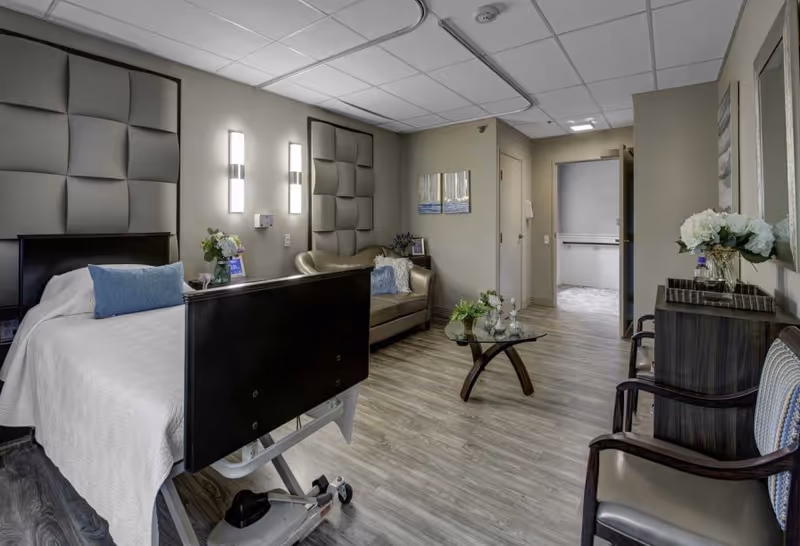 A well-lit senior living bedroom featuring a hospital-style bed with white bedding and a blue pillow, a small beige loveseat with a decorative pillow, a glass coffee table with flowers, and two chairs with cushions. The room has wood flooring, padded wall panels behind the bed, modern wall lights, and a doorway leading to another room.