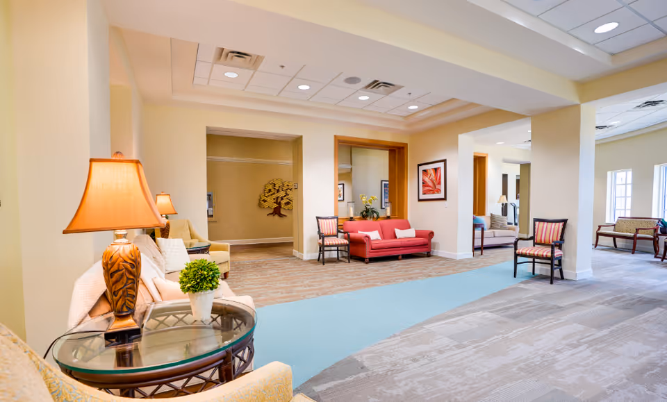 A spacious and well-lit senior living facility common area with various seating options including armchairs, a red sofa, and wooden chairs. The room features beige walls, a mix of carpet and wood flooring, decorative lamps, framed artwork, and a small potted plant on a glass-top side table.
