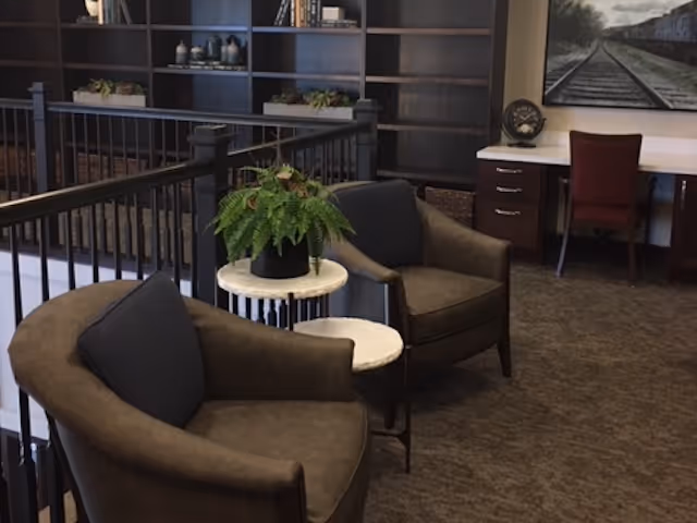 Two upholstered armchairs with small marble-topped side tables and a potted fern in a carpeted lounge area with shelving and a desk in the background.