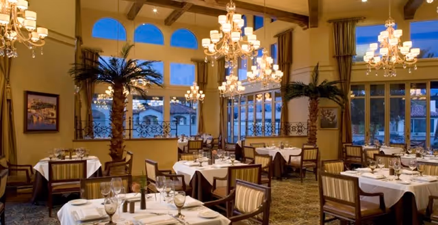 Elegant dining room with multiple tables covered in white tablecloths, set with glassware and silverware. The room features large windows with views of an outdoor area, tall palm plants, chandeliers hanging from a high ceiling with exposed wooden beams, and striped upholstered chairs.
