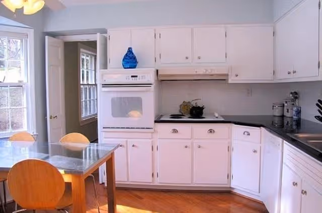 A bright kitchen with white cabinets, a built-in oven, a stovetop, and a black countertop. There is a glass dining table with wooden chairs in the foreground, and sunlight is streaming in through the windows.