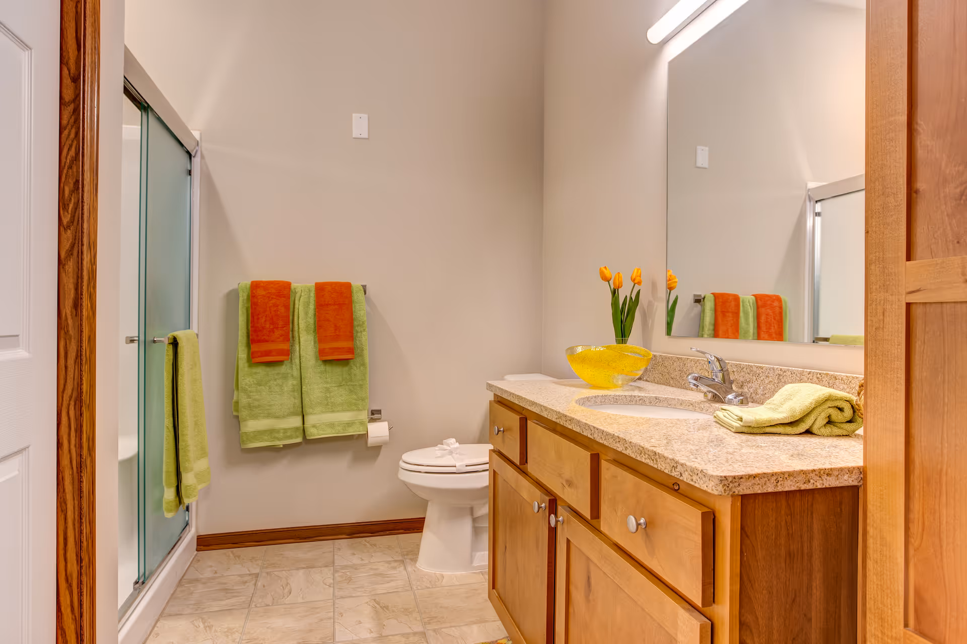 A clean bathroom with a wooden vanity topped with a granite countertop and a sink. There is a large mirror above the vanity, a toilet with a folded towel on the seat, and a glass-enclosed shower. Two green towels with orange towels on top hang on a wall-mounted towel rack, and a green towel hangs on the shower door. A yellow glass bowl and a vase with orange tulips are on the countertop.