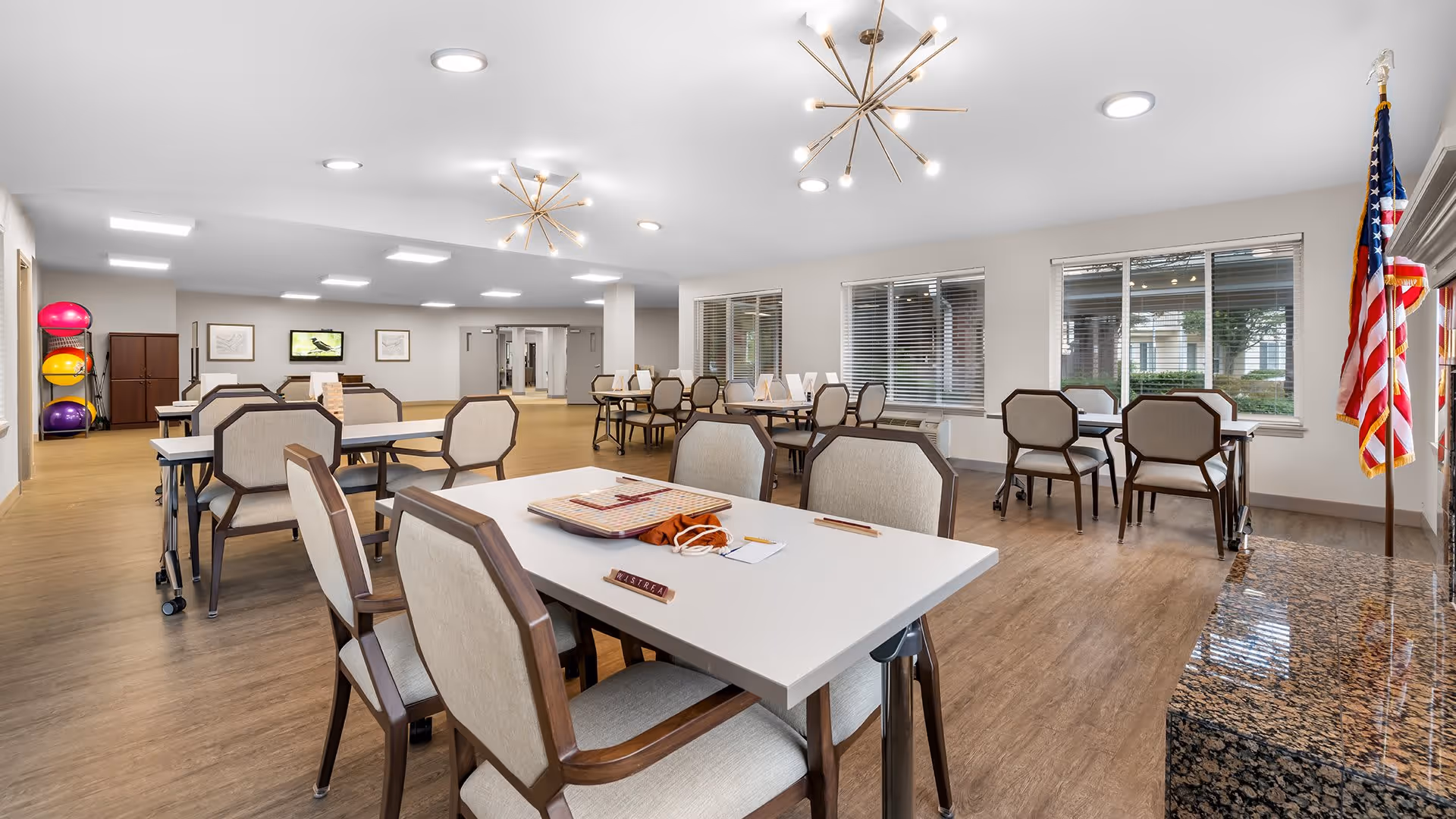 A spacious, well-lit common area with multiple tables and chairs arranged for group activities or dining. The room features large windows with blinds, modern ceiling lights, and an American flag near a granite countertop. Exercise balls are stacked on a rack in the corner, and a television is mounted on the far wall.