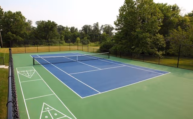 Outdoor tennis court with a blue playing surface, green outer area, net and surrounding fence with trees in the background.