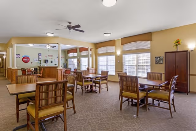 A spacious dining room with multiple wooden tables and chairs arranged neatly. The room has large windows with blinds allowing natural light to fill the space. The walls are painted a warm beige color, and there are ceiling fans and wall-mounted lights providing illumination. In the background, there is a counter area with a Coca-Cola sign and various decorations on the wall.