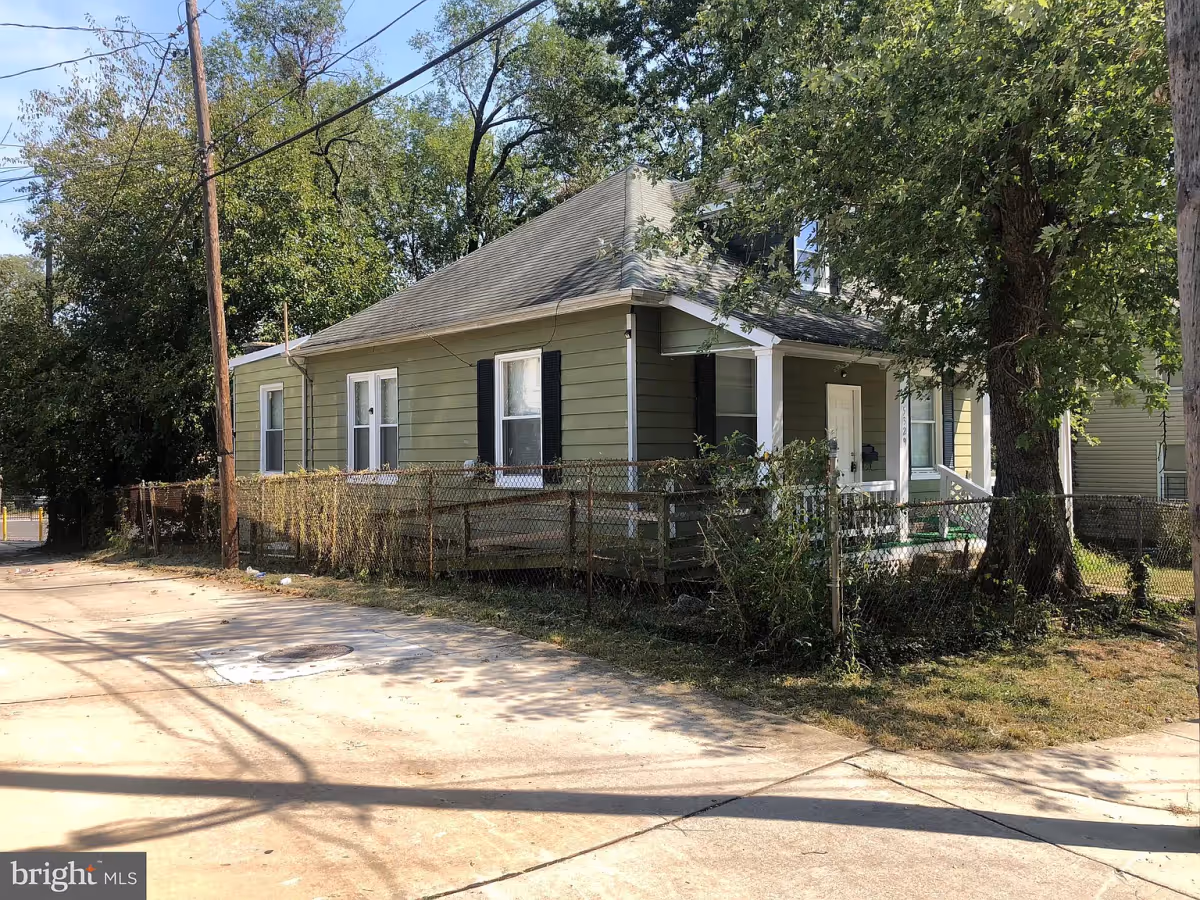 Exterior view of a single-story green house with white trim and black shutters, surrounded by a chain-link fence and trees, under a clear sky.