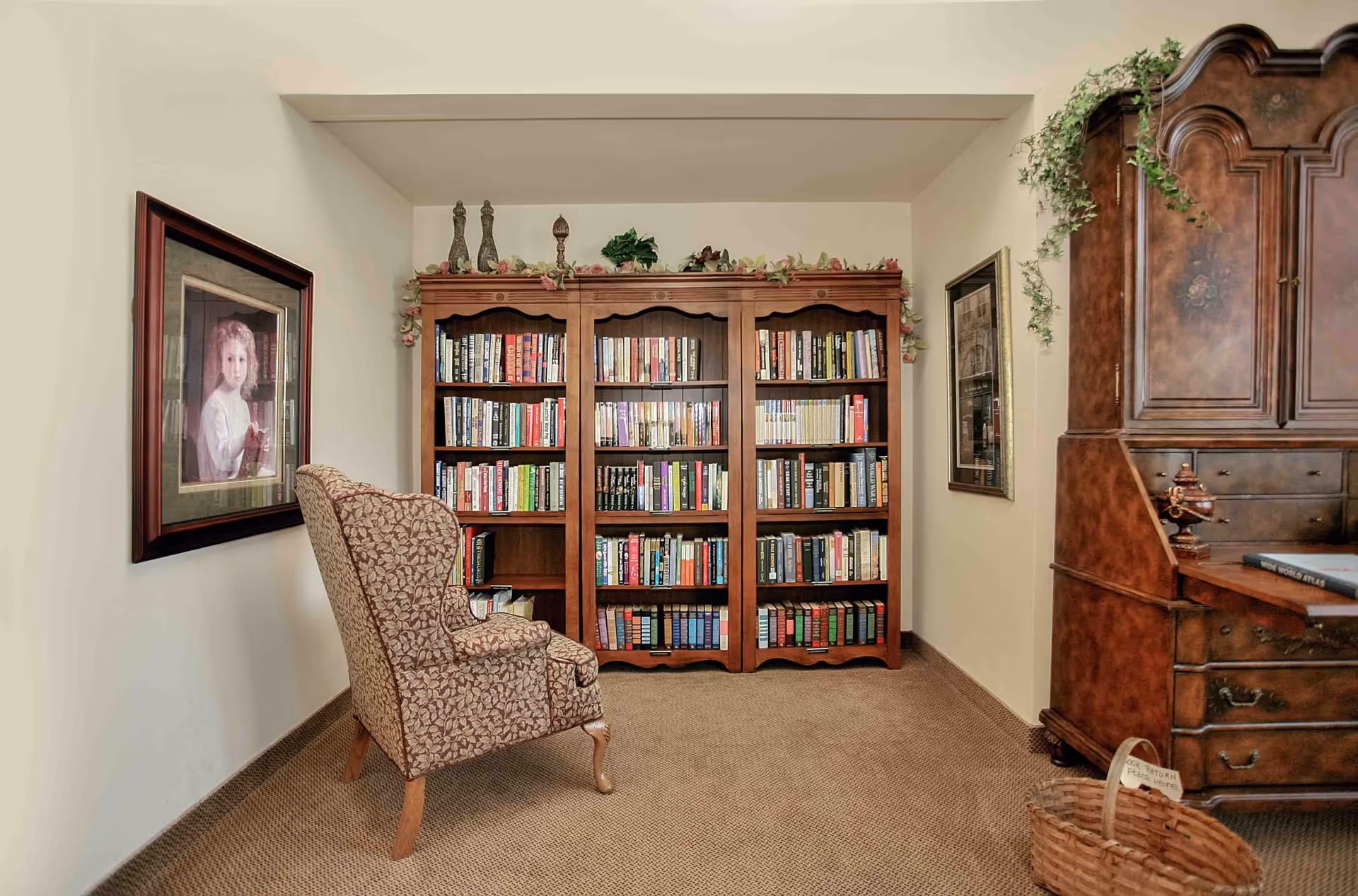 A cozy reading nook featuring a patterned armchair facing a large wooden bookshelf filled with books. The bookshelf is decorated with greenery and small ornamental items on top. To the right is a wooden antique-style cabinet with a plant draping over it. On the left wall, there is a framed portrait of a young child. The room has beige carpet and light-colored walls.