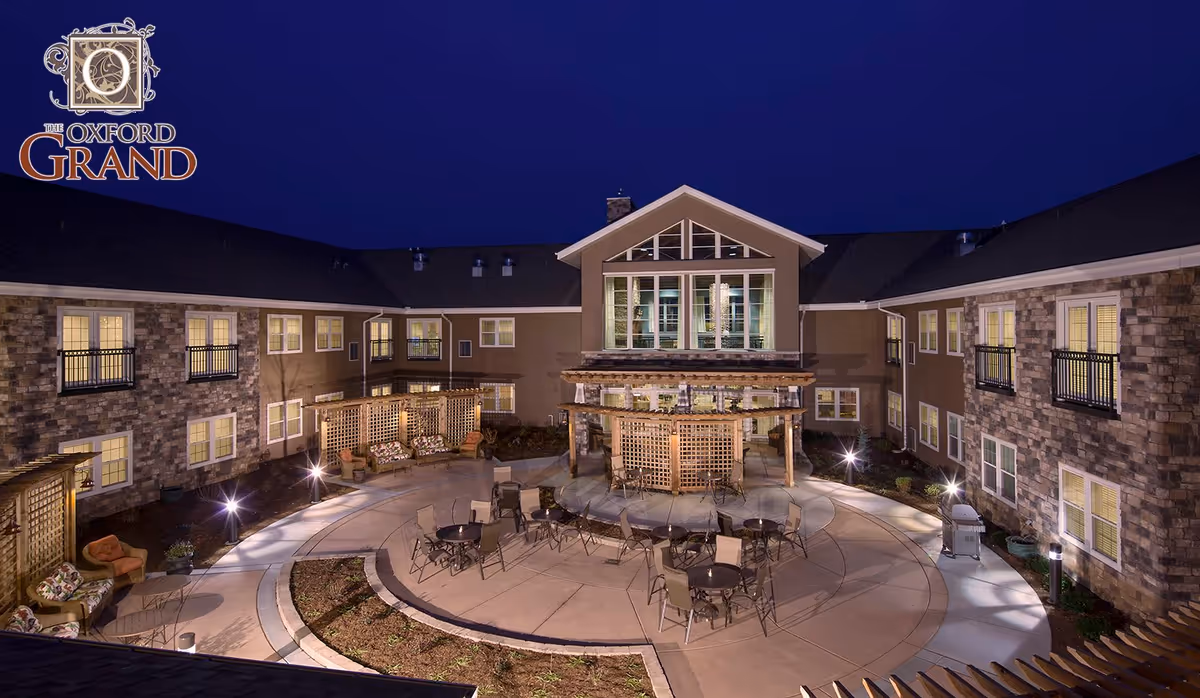 Night view of the outdoor courtyard at The Oxford Grand Assisted Living & Memory Care, featuring a circular paved area with tables and chairs, surrounded by seating areas with cushioned chairs and lattice structures. The building has stone and brown exterior walls with many lit windows.