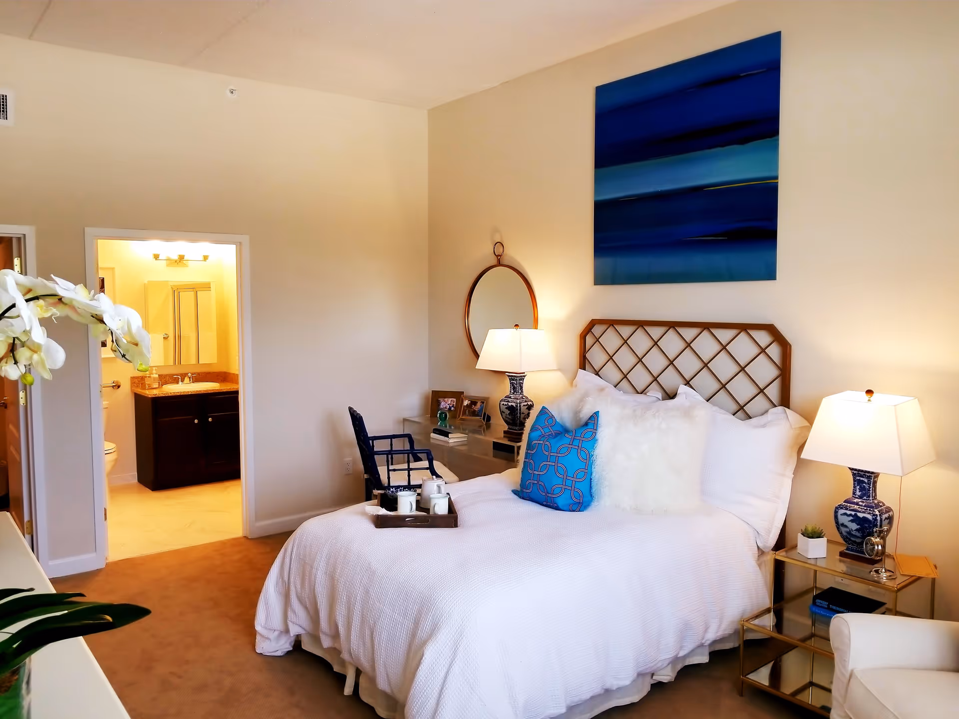 A cozy bedroom with a neatly made bed featuring white bedding and decorative pillows, a blue patterned pillow, and a lattice-style headboard. There are two bedside tables with matching blue and white lamps, a round mirror on the wall, a chair, and a small desk with books and framed photos. An open door reveals a bathroom with a sink, mirror, and shower. A white orchid plant is partially visible in the foreground.