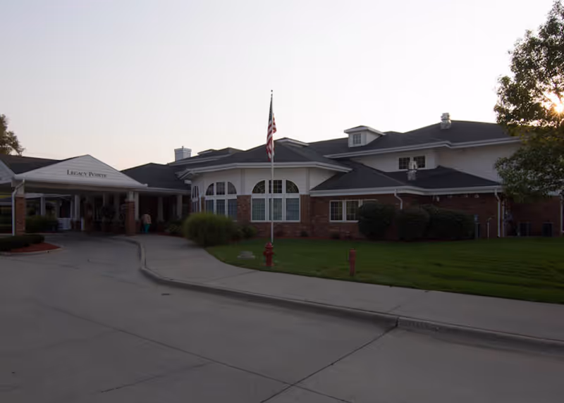 Exterior view of a senior living facility building with a driveway, an American flag on a flagpole, and well-maintained green lawn and bushes. The building has a brick and white facade with multiple windows and a covered entrance labeled 'Legacy Pointe'.
