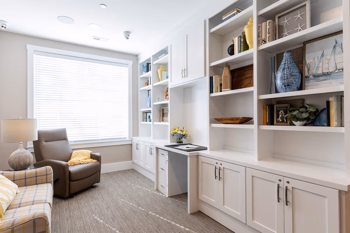 Bright sitting room with built-in white bookshelves, a recliner and a plaid sofa by a large window with blinds.
