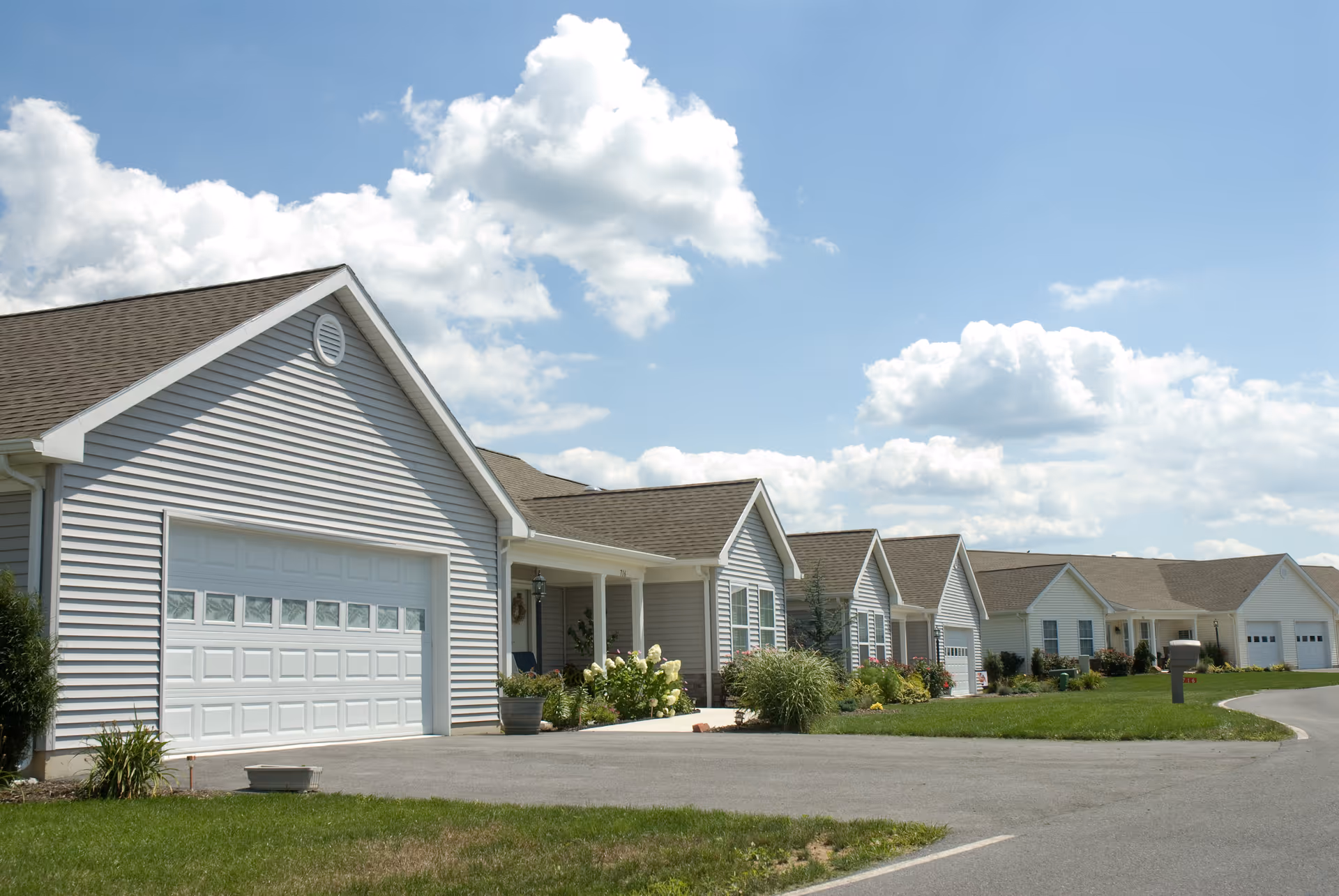 A row of single-story residential buildings with garages, driveways, and well-maintained lawns under a partly cloudy blue sky.