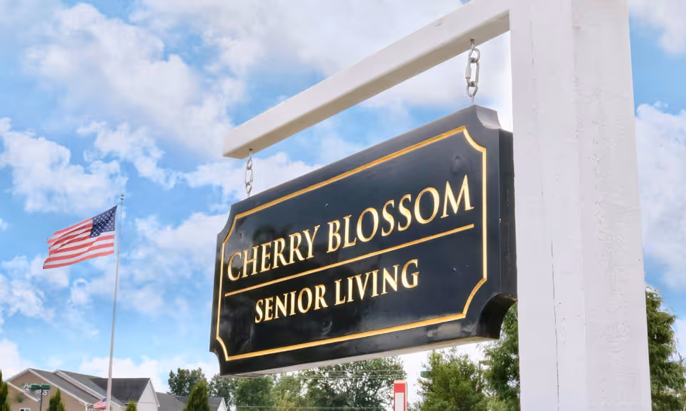 A hanging sign reading Cherry Blossom Senior Living outdoors with an American flag nearby.