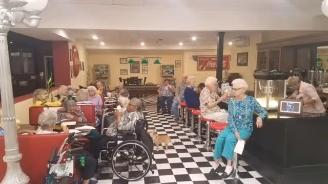 A group of elderly people sitting and socializing in a retro-style dining area with black and white checkered floor tiles, red stools at a counter, and red booth seating. Some individuals are in wheelchairs, and a small dog is walking on the floor. The room is decorated with vintage Coca-Cola signs and framed pictures on the walls.