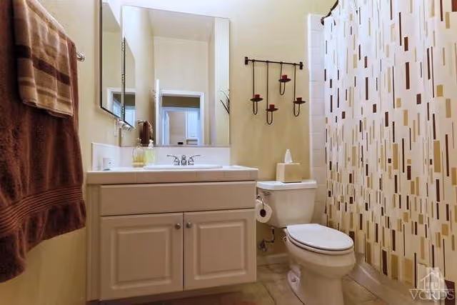 A clean bathroom featuring a white vanity with a sink and faucet, a large mirror above the vanity, a toilet with a tissue box on top, a wall-mounted candle holder with three candles, a shower with a patterned curtain, and a brown towel hanging on a towel rack.