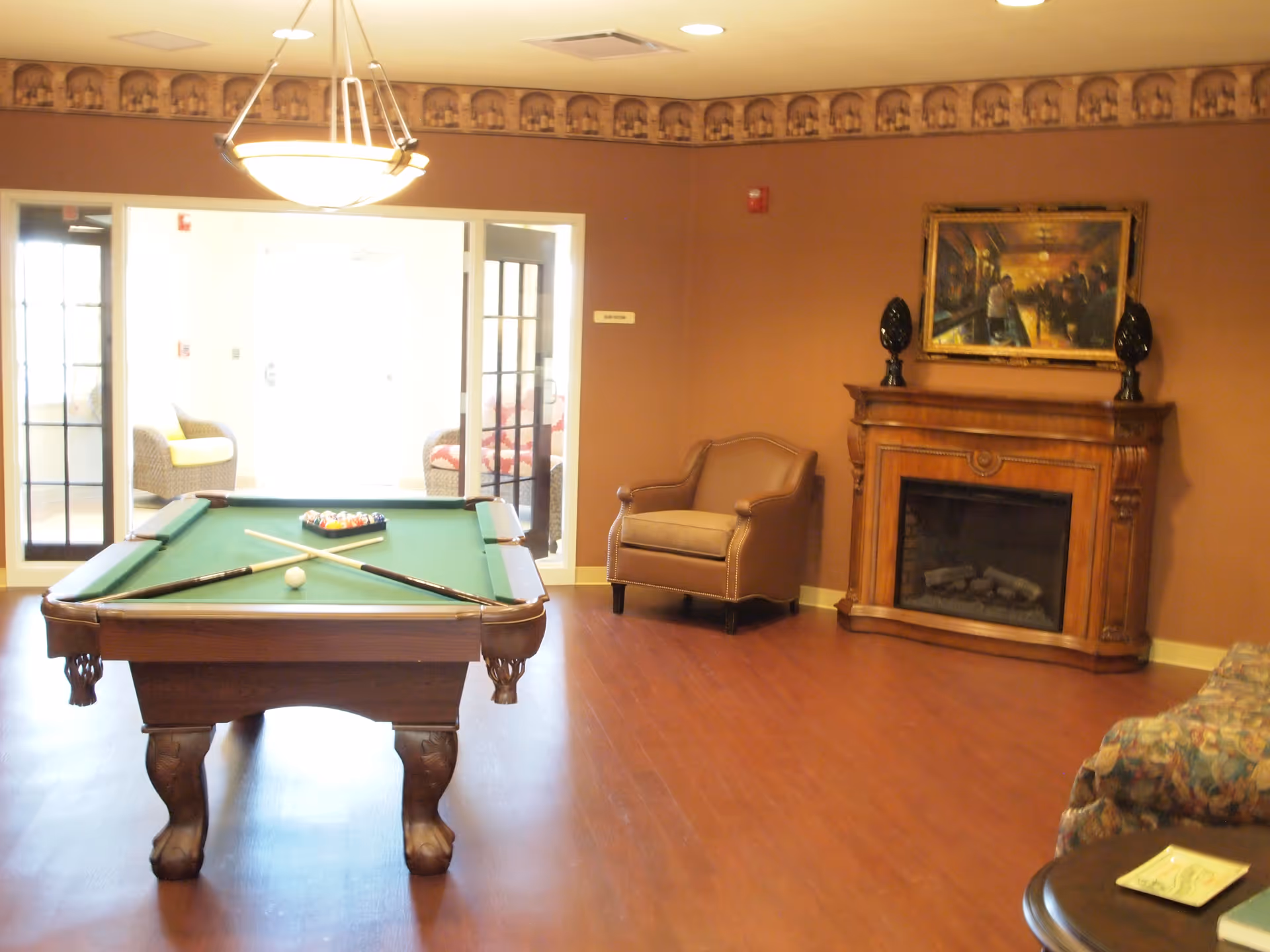 A cozy lounge with a pool table in the center, an armchair, and a decorative fireplace topped by a painting.