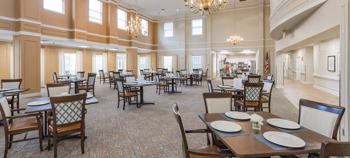 A spacious dining room with multiple wooden tables and chairs arranged neatly. Each table is set with white plates and placemats. The room features high ceilings with large windows allowing natural light to fill the space. Elegant chandeliers hang from the ceiling, and the walls are painted in a warm beige tone with white trim. There is an American flag visible in the background near the kitchen area.