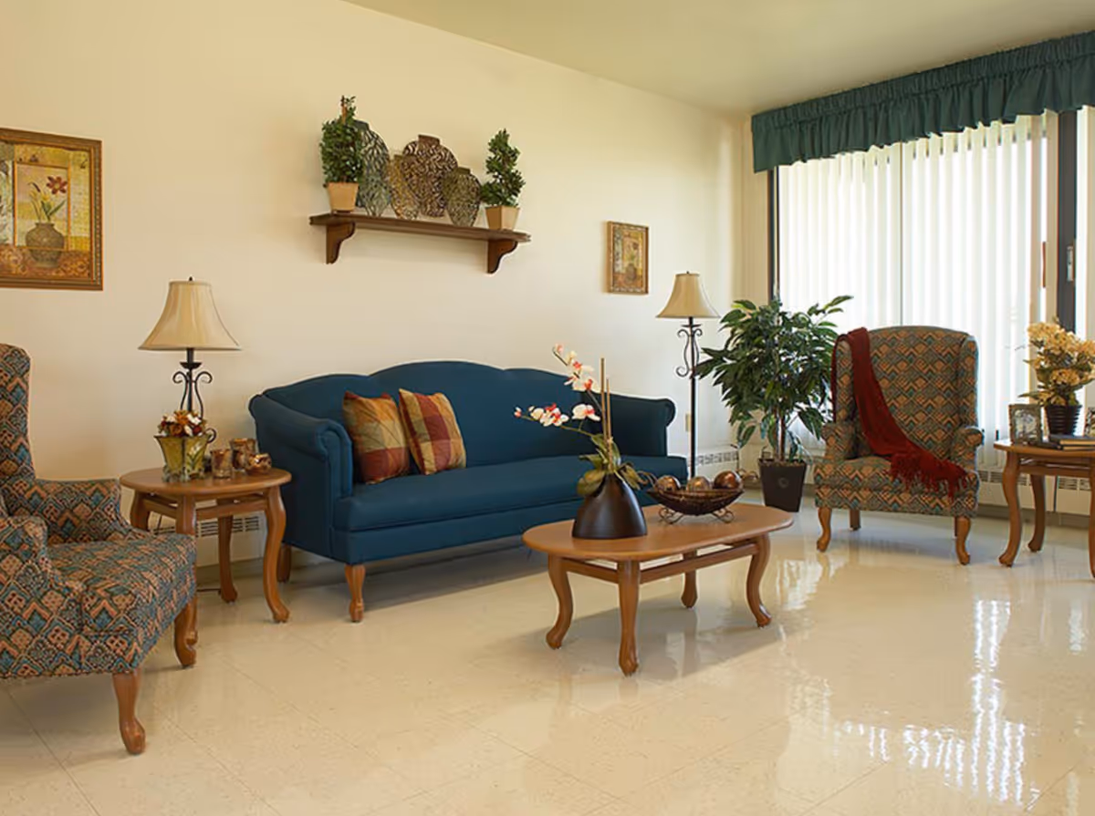 A cozy living room with a blue upholstered sofa adorned with patterned cushions, two patterned armchairs, wooden coffee and side tables with decorative items, two table lamps, potted plants, framed artwork on the walls, and large vertical blinds covering a window.