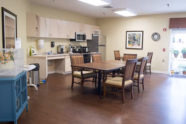 A spacious dining area with a wooden table and six chairs with cushioned seats. In the background, there is a kitchenette with light wood cabinets, a microwave, a coffee maker, and a refrigerator. The room has light-colored walls, a framed picture, a clock, and a window with potted plants on the sill. The floor is a dark wood laminate.
